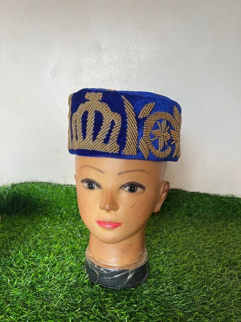 May include: A blue velvet hat with gold embroidered designs. The hat is placed on a mannequin head with a neutral skin tone. The hat's design features geometric and stylized floral patterns. The background is a mix of green and white.