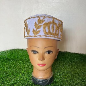 May include: A white and gold embroidered hat with a decorative design. The hat is displayed on a mannequin head with a neutral skin tone. The hat features gold embroidery around the brim and a floral pattern.