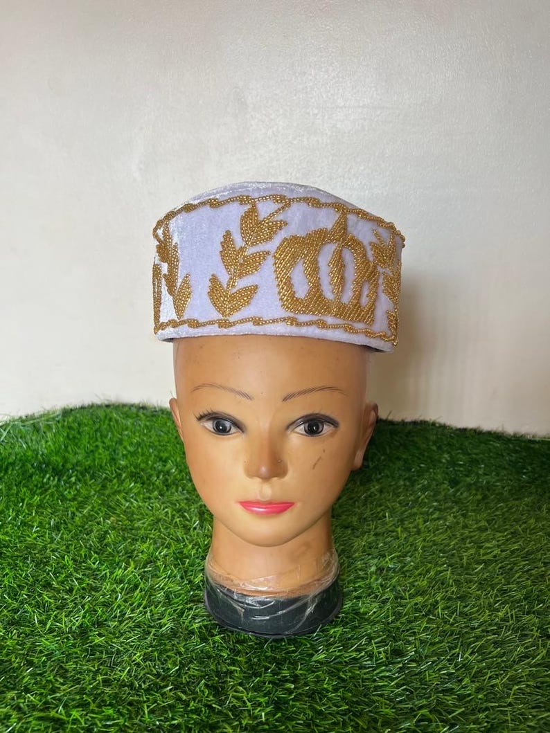 May include: A white and gold embroidered hat with a decorative design. The hat is displayed on a mannequin head with a neutral skin tone. The hat features gold embroidery around the brim and a floral pattern.