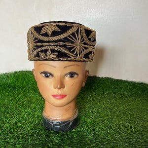 May include: A black velvet hat with gold embroidery. The hat is rectangular with a flat top and a band of gold embroidery around the base. The gold embroidery features a floral design. The hat is displayed on a mannequin head.