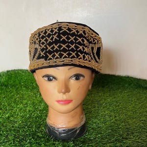 May include: A black hat with gold-colored beaded embroidery. The hat is displayed on a mannequin head with a neutral skin tone. The embroidery forms a geometric pattern across the top and sides of the hat. The background is a light green surface.