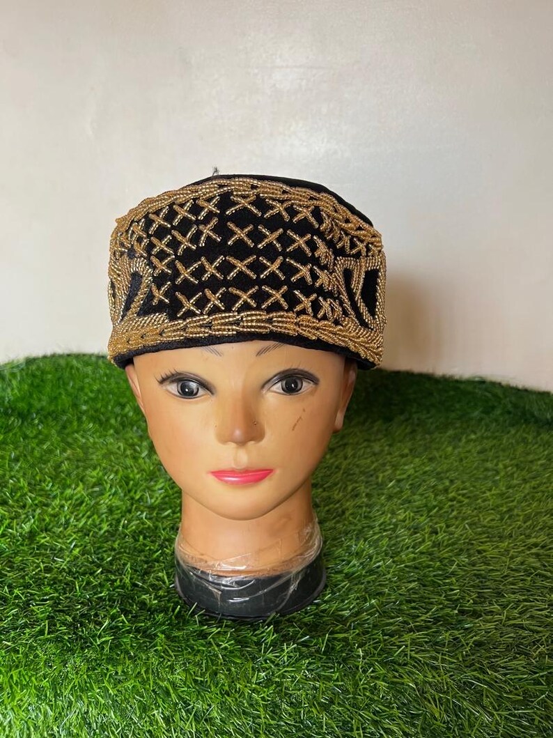 May include: A black hat with gold-colored beaded embroidery. The hat is displayed on a mannequin head with a neutral skin tone. The embroidery forms a geometric pattern across the top and sides of the hat. The background is a light green surface.
