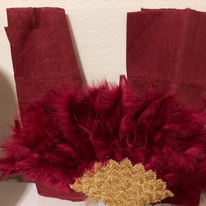 May include: A burgundy feather fan with a gold beaded center. The fan is surrounded by two burgundy fabric pieces.