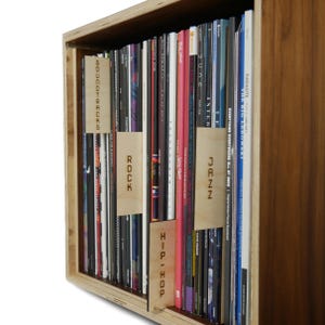 Puede incluir: Una estantería de madera con discos de vinilo organizados por género. Los géneros están etiquetados con divisores de madera: Bandas sonoras, Rock, Jazz y Hip-Hop.