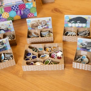 May include: A collection of cat-themed game pieces and cards. The cards feature illustrations of cats, and the game pieces are colorful, decorative tokens. The items are arranged in small, divided cardboard boxes, with a larger box in the background.
