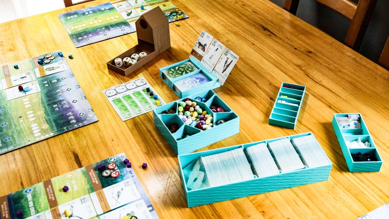 May include: A board game setup featuring a wooden dice tower, several light blue plastic organizers containing decks of cards, small colorful tokens, and game boards with green and white illustrations spread across a wooden table surface.