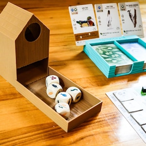May include: A tan 3D-printed birdhouse-shaped dice tower with a tray containing five white dice with colored icons. Beside it sits a light blue plastic card holder with two decks of cards, three upright bird cards, and a game board with round markers.