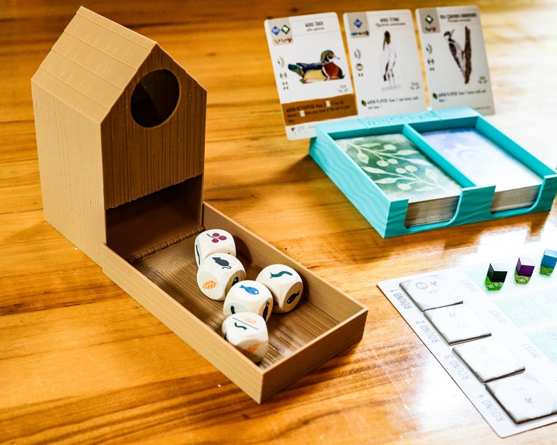 May include: A tan 3D-printed birdhouse-shaped dice tower with a tray containing five white dice with colored icons. Beside it sits a light blue plastic card holder with two decks of cards, three upright bird cards, and a game board with round markers.