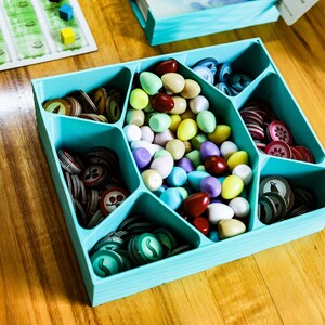 May include: A light blue plastic board game organizer tray with six compartments. The central section holds a collection of small, smooth, egg-shaped tokens in various colors, while the surrounding sections contain stacks of circular cardboard game tokens.