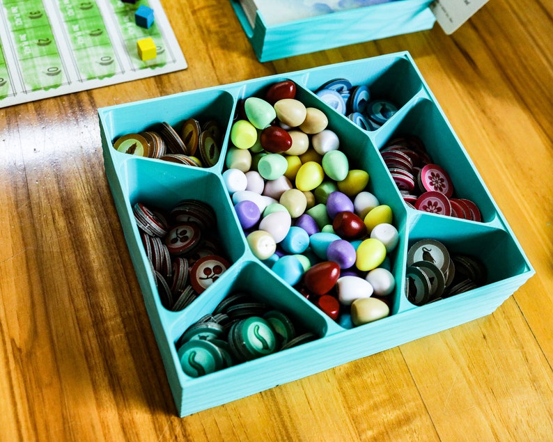 May include: A light blue plastic board game organizer tray with six compartments. The central section holds a collection of small, smooth, egg-shaped tokens in various colors, while the surrounding sections contain stacks of circular cardboard game tokens.