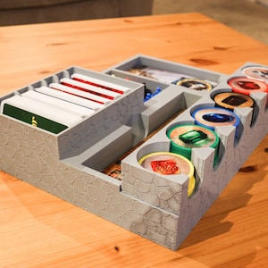 May include: A gray 3D printed organizer for a board game. The organizer has compartments for cards, tokens, and dice. The compartments are labeled with the names of the game components.