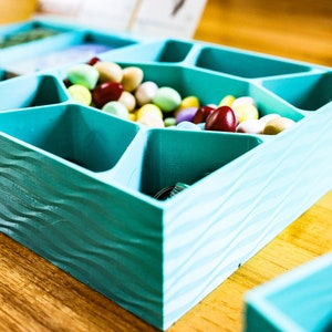 May include: A square, teal-colored storage tray with a wavy, textured exterior and multiple internal compartments. The tray is filled with a variety of small, smooth, multi-colored oval candies in shades of yellow, pink, white, and dark red.