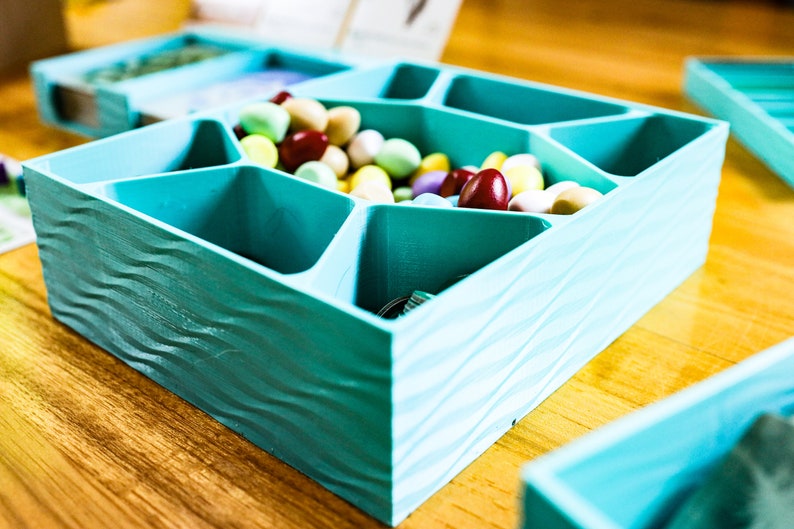 May include: A square, teal-colored storage tray with a wavy, textured exterior and multiple internal compartments. The tray is filled with a variety of small, smooth, multi-colored oval candies in shades of yellow, pink, white, and dark red.