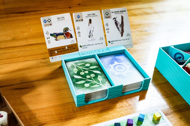 May include: A light blue plastic card holder for the board game Wingspan, featuring a central divider and a slot for three upright game cards. The cards display illustrations of a Wood Duck, a Wood Stork, and a Red-Cockaded Woodpecker.