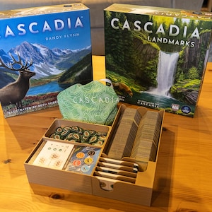 May include: Two board game boxes titled Cascadia and Cascadia Landmarks, a light green fabric drawstring bag with the word Cascadia printed on it, and a tan plastic organizer tray containing stacks of cards, game tokens, and small tiles.