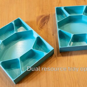 May include: Two rectangular turquoise plastic trays, each featuring a central hexagonal compartment surrounded by six smaller, irregularly shaped sections. The text "Dual resource tray option" is printed in white at the bottom center.