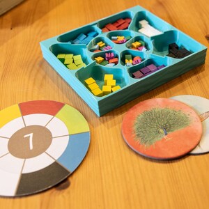 May include: A teal plastic organizer tray filled with small wooden cubes in red, white, black, blue, green, yellow, and purple. Beside the tray are two circular cardboard game tokens, one featuring a color wheel with the number 7 and one with a peacock.