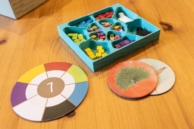 May include: A teal plastic organizer tray filled with small wooden cubes in red, white, black, blue, green, yellow, and purple. Beside the tray are two circular cardboard game tokens, one featuring a color wheel with the number 7 and one with a peacock.
