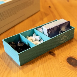 May include: A rectangular teal board game organizer with three internal compartments. The left section holds black tokens, the middle section contains white tokens, and the right section stores a deck of playing cards with a dark blue graphic design.