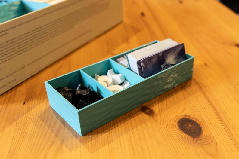 May include: A rectangular teal board game organizer with three internal compartments. The left section holds black tokens, the middle section contains white tokens, and the right section stores a deck of playing cards with a dark blue graphic design.