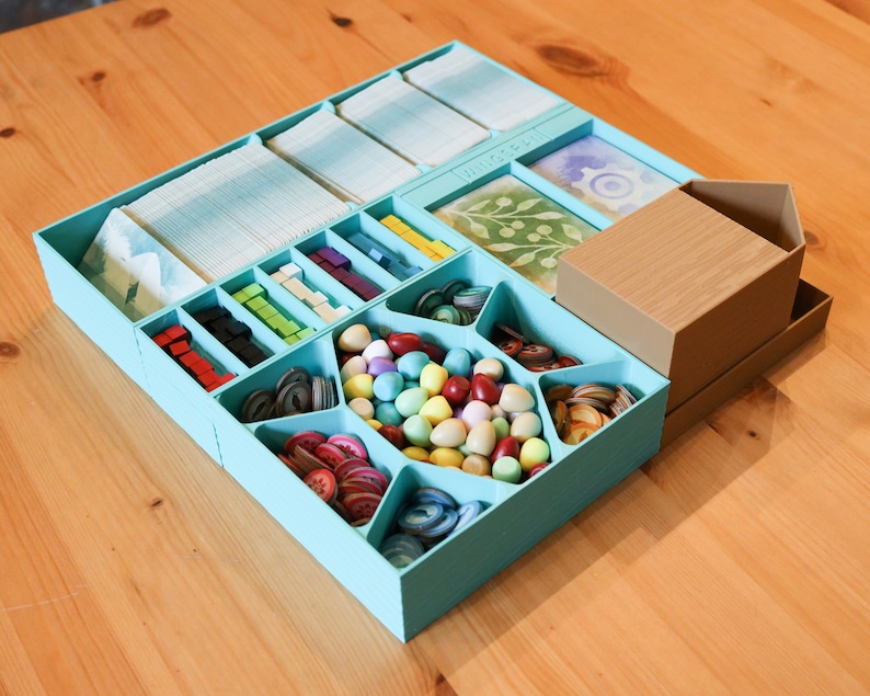 May include: A light blue plastic board game organizer tray with multiple compartments containing various game components, including stacks of cards, small colored cubes, circular tokens, and a collection of pastel egg-shaped pieces, with a brown house-shaped box.