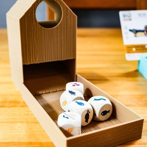 May include: A tan, 3D-printed plastic dice tower shaped like a small house with a circular opening at the top. Inside the attached tray are five white wooden dice, each featuring various black, blue, orange, and purple icons of animals and plants.