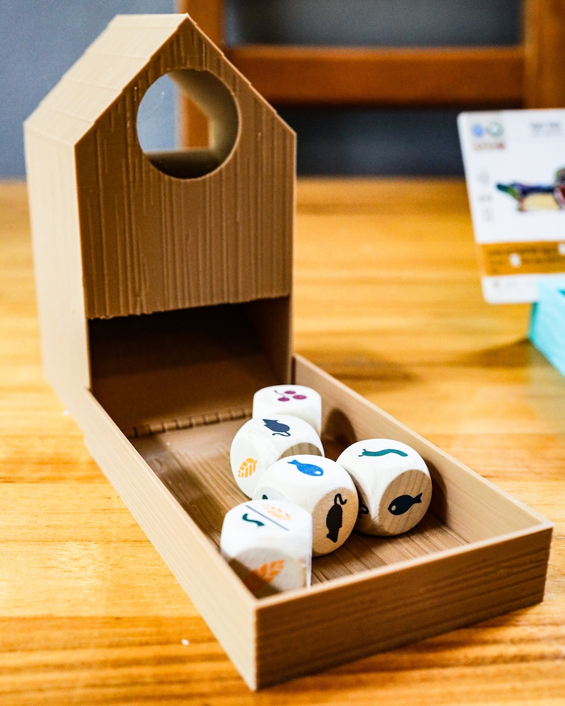 May include: A tan, 3D-printed plastic dice tower shaped like a small house with a circular opening at the top. Inside the attached tray are five white wooden dice, each featuring various black, blue, orange, and purple icons of animals and plants.