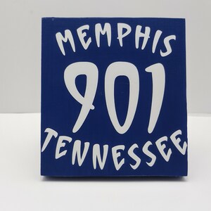 May include: A blue wooden block with white lettering. The text reads "MEMPHIS 901 TENNESSEE". The block is a square shape and is likely a decorative item or souvenir.