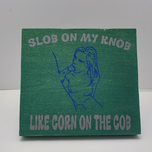 May include: A green wooden block with the text "SLOB ON MY KNOB" above a blue line drawing of a person and the text "LIKE CORN ON THE COB" below.
