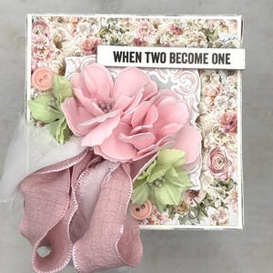 May include: A square box decorated with a floral pattern, a large pink flower, and green leaves. A pink ribbon and two buttons are attached. The text "WHEN TWO BECOME ONE" is on a white banner.