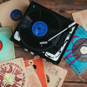 May include: A vintage record player with a black turntable and a silver tone arm. The record player is surrounded by various vinyl records with colorful covers. The records include titles like "Parlez Avec" and "Nagrania Polskie Nagrania".