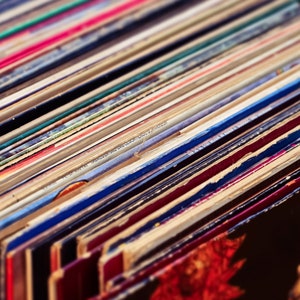 May include: A stack of vintage vinyl records in various colors, including red, blue, green, and yellow. The records are stacked in a neat pile, with the edges of the records visible.