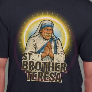 May include: Navy blue t-shirt featuring an illustration of Mother Teresa with the text "ST. BROTHER TERESA". The graphic shows Mother Teresa with her hands clasped in prayer, surrounded by a golden halo.