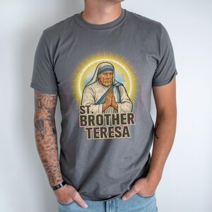 May include: Gray t-shirt featuring a graphic of St. Brother Teresa with a halo, hands clasped in prayer, and the text "ST. BROTHER TERESA." The shirt is worn by a person with tattoos on their arm.