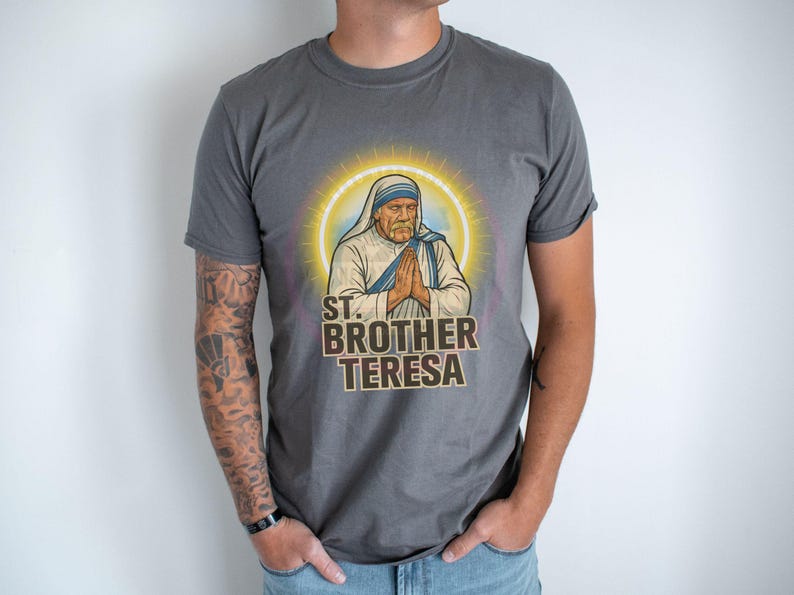 May include: Gray t-shirt featuring a graphic of St. Brother Teresa with a halo, hands clasped in prayer, and the text "ST. BROTHER TERESA." The shirt is worn by a person with tattoos on their arm.