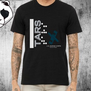 Op de afbeelding: Zwart T-shirt met een witte afbeelding van de tekst "TARS" en een gestileerde afbeelding van een robot met de tekst "U.S. MARINE CORPS Tactical Robot".