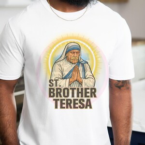 May include: White t-shirt featuring a graphic of St. Brother Teresa with hands clasped in prayer. The design includes a halo and the text "ST. BROTHER TERESA" in bold letters. The shirt is made of a soft material.