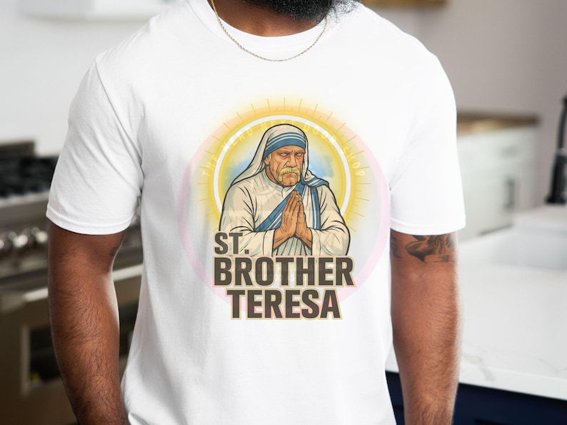 May include: White t-shirt featuring a graphic of St. Brother Teresa with hands clasped in prayer. The design includes a halo and the text "ST. BROTHER TERESA" in bold letters. The shirt is made of a soft material.