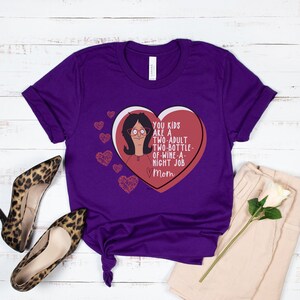 K&ouml;nnte beinhalten: Ein lila T-Shirt mit einem Cartoon-Bild einer Frau in einem herzf&ouml;rmigen Bonbon. Das Bonbonherz tr&auml;gt den Text "YOU KIDS ARE A TWO-ADULT TWO-BOTTLE-OF-WINE-A-NIGHT JOB Mom". Mehrere kleine Herzgrafiken befinden sich ebenfalls auf dem T-Shirt.