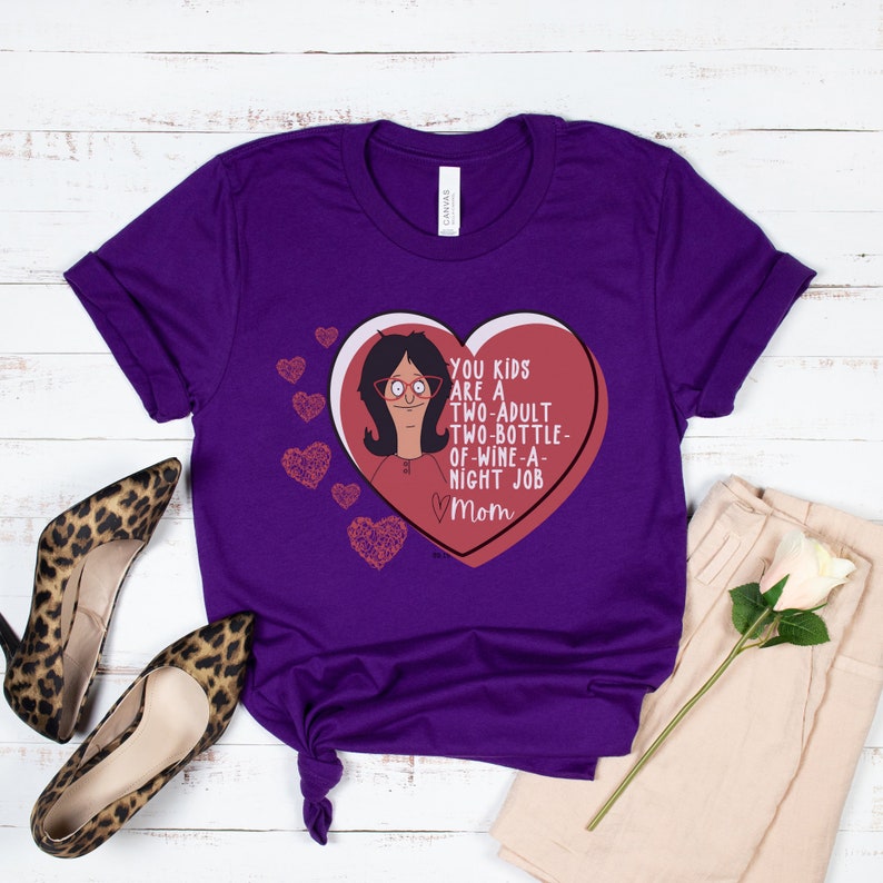 K&ouml;nnte beinhalten: Ein lila T-Shirt mit einem Cartoon-Bild einer Frau in einem herzf&ouml;rmigen Bonbon. Das Bonbonherz tr&auml;gt den Text "YOU KIDS ARE A TWO-ADULT TWO-BOTTLE-OF-WINE-A-NIGHT JOB Mom". Mehrere kleine Herzgrafiken befinden sich ebenfalls auf dem T-Shirt.