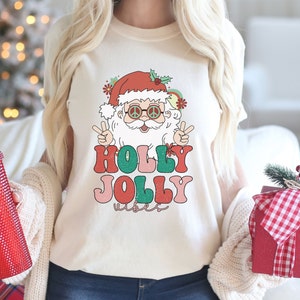 May include: A white t-shirt with a retro-style illustration of Santa Claus wearing sunglasses and peace signs. The text "HOLLY JOLLY vibes" is printed below the illustration in colorful, vintage-style lettering.