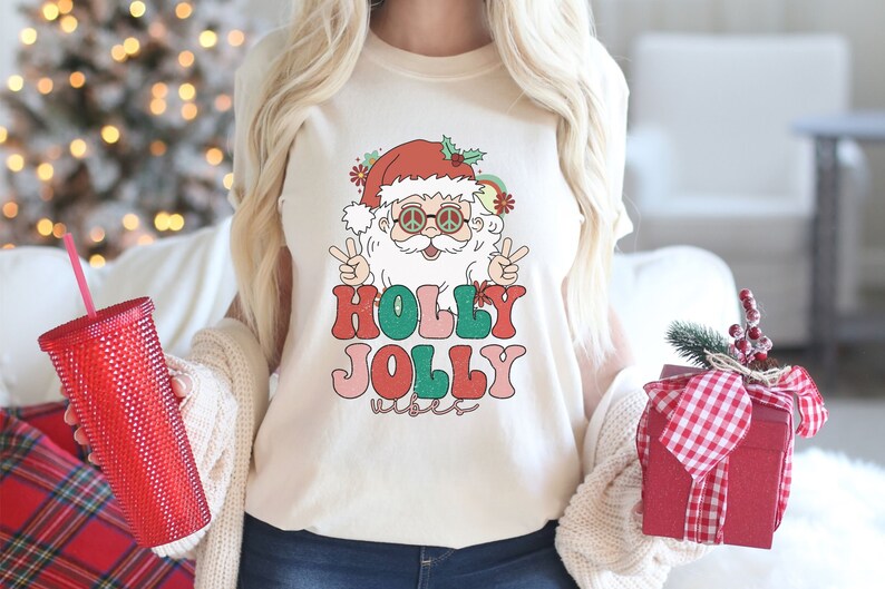May include: A white t-shirt with a retro-style illustration of Santa Claus wearing sunglasses and peace signs. The text "HOLLY JOLLY vibes" is printed below the illustration in colorful, vintage-style lettering.
