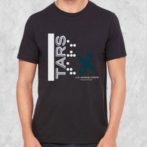 Op de afbeelding: Zwart T-shirt met een wit grafisch ontwerp met de tekst "TARS" met een reeks stippen en een gestileerd robotontwerp. De tekst onder het ontwerp luidt "U.S. MARINE CORPS Tactical Robot".