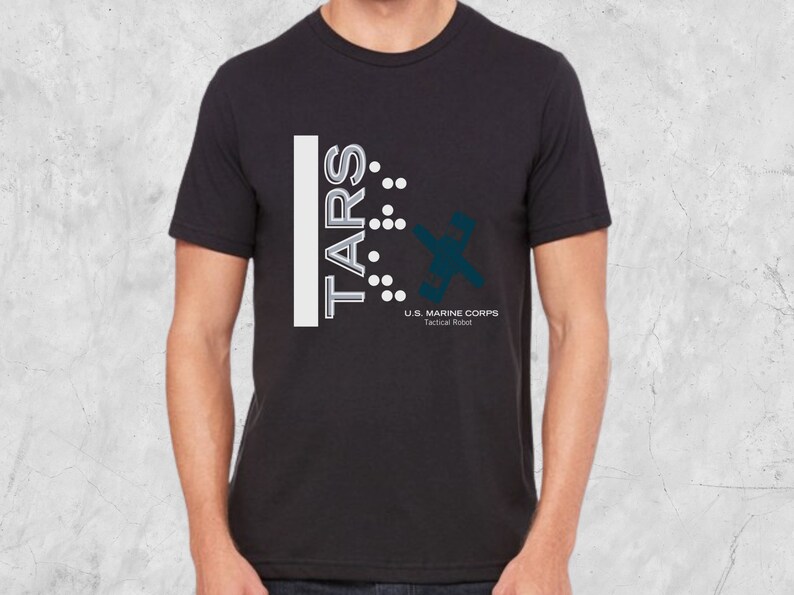 Op de afbeelding: Zwart T-shirt met een wit grafisch ontwerp met de tekst "TARS" met een reeks stippen en een gestileerd robotontwerp. De tekst onder het ontwerp luidt "U.S. MARINE CORPS Tactical Robot".