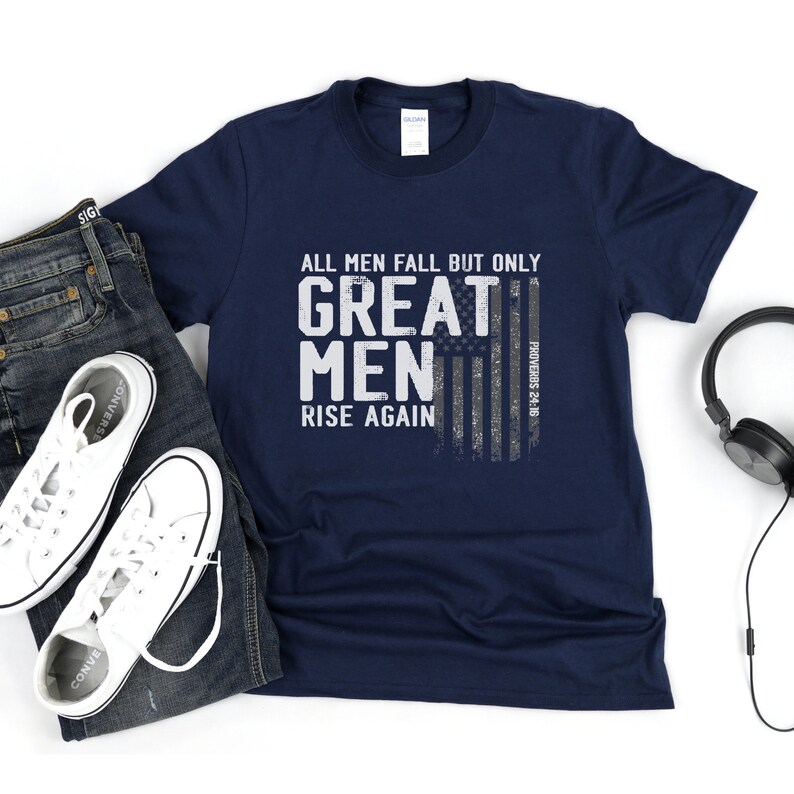 Puede incluir: Una camiseta azul marino con un gr&aacute;fico blanco desgastado que dice "All men fall but only great men rise again" con un dise&ntilde;o de bandera estadounidense descolorido y la escritura "Proverbios 24:16".
