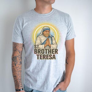 May include: A light gray t-shirt featuring a graphic of St. Brother Teresa with a halo. The image shows St. Brother Teresa with hands clasped in prayer. The text "ST. BROTHER TERESA" is printed below the image in brown.