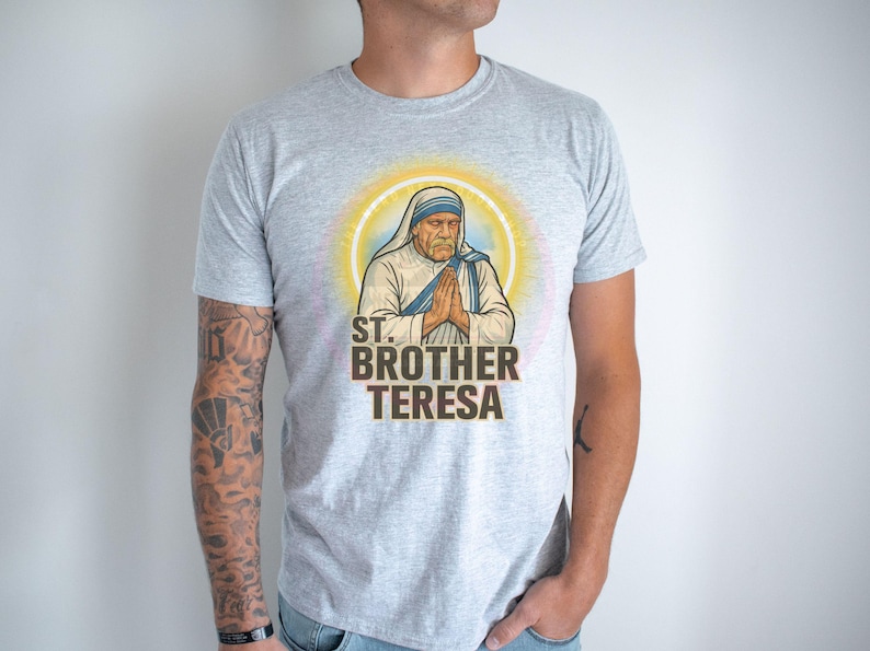 May include: A light gray t-shirt featuring a graphic of St. Brother Teresa with a halo. The image shows St. Brother Teresa with hands clasped in prayer. The text "ST. BROTHER TERESA" is printed below the image in brown.