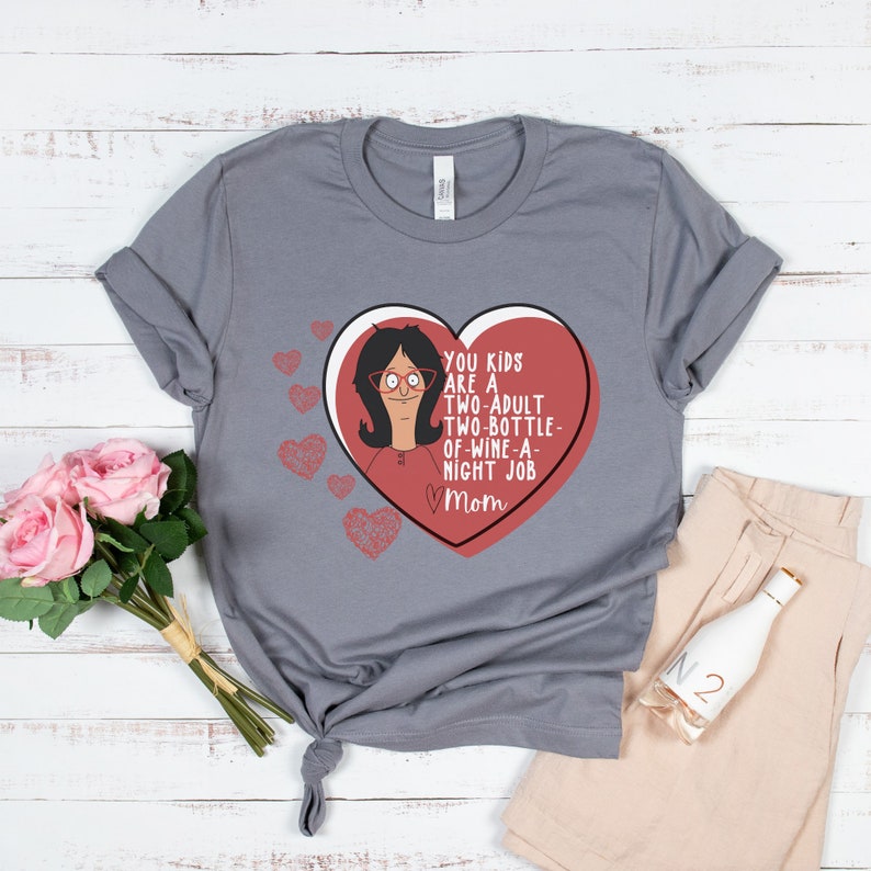 K&ouml;nnte beinhalten: Graues T-Shirt mit herzf&ouml;rmigem Design, Cartoon-Figur und dem Text "YOU KIDS ARE A TWO-ADULT TWO-BOTTLE-OF-WINE-A-NIGHT JOB Mom". Das Bild zeigt beige Shorts und eine dekorative Flasche.