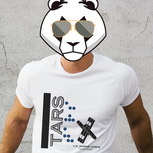 Op de afbeelding: Wit T-shirt met een zwart en grijs grafisch ontwerp met de tekst "TARS" met blauwe stippen en de tekst "U.S. MARINE CORPS Tactical Robot".