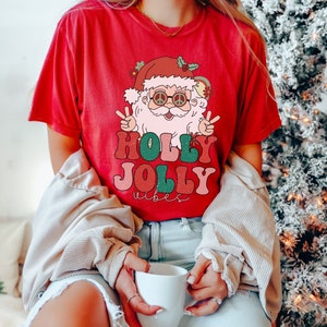 May include: Red t-shirt with a retro-style illustration of Santa Claus wearing sunglasses and peace signs. The text "HOLLY JOLLY vibes" is printed in colorful letters.